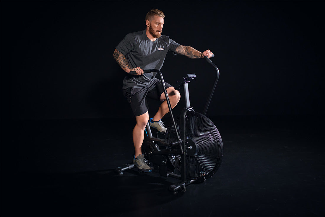A man with tattoos, wearing a gray t-shirt, black shorts, and sneakers, exercises on an air bike against a dark background. He is gripping the handles and pedaling, focused on his workout.