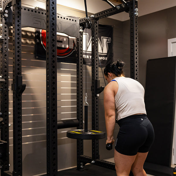 Wearing athletic wear, a person in a home gym prepares for functional strength training with the SMAI Cable Pulley System, gripping its handle attached to the round weight platform.