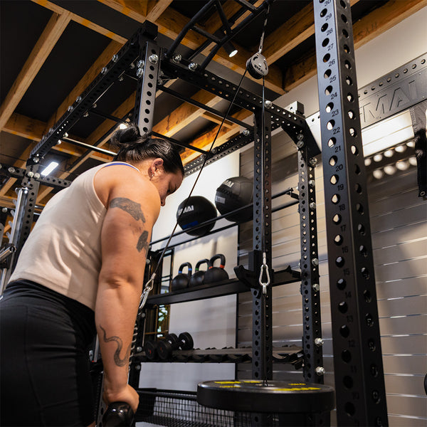 A tattooed individual uses the SMAI Cable Pulley System for functional strength training in a gym, pulling down the bar with both hands. Various weights and other gym equipment appear in the background.