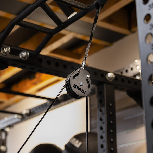 Close-up of the SMAI Cable Pulley System in black, attached to a metal power rack—ideal for functional strength training at home. Black cables run through the pulley with an exposed wooden ceiling in the background.