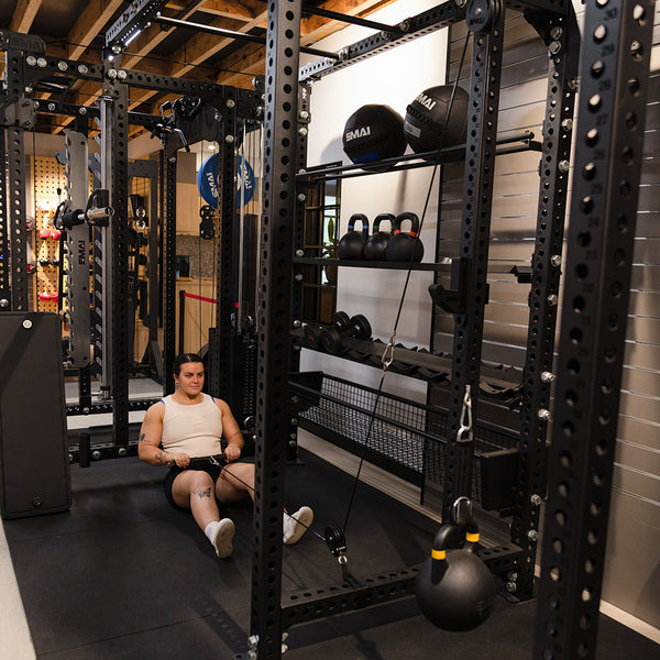 A person uses the SMAI Cable Pulley System for seated rows on a gym floor, with kettlebells, medicine balls, and other functional strength equipment in the background.