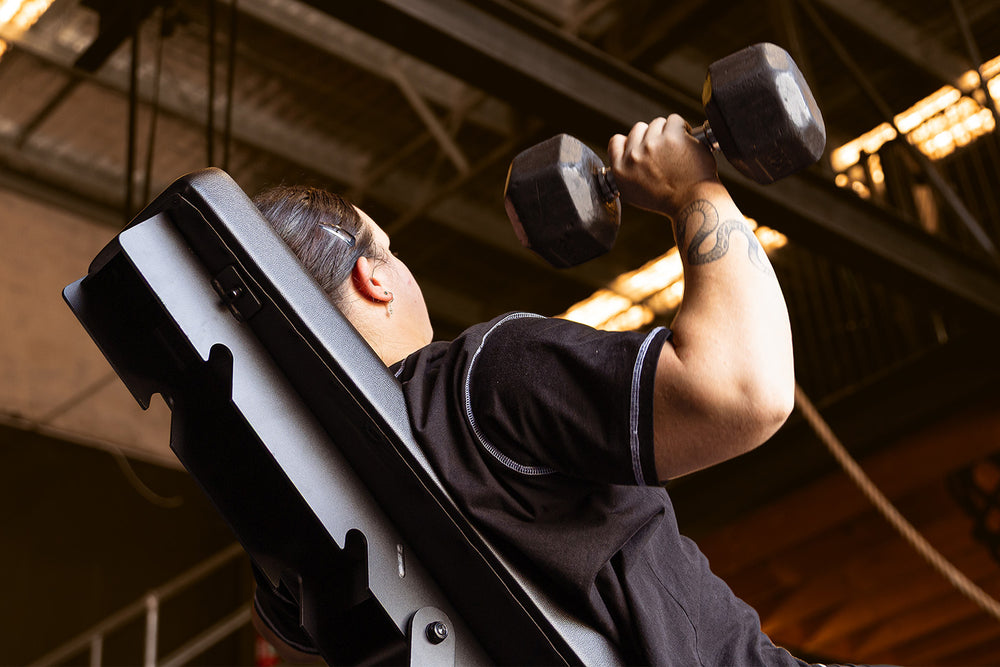 A person with a tattoo on their arm is lying on an incline bench in a gym, lifting a dumbbell overhead with one hand. The gym has metal beams and industrial lighting in the background.