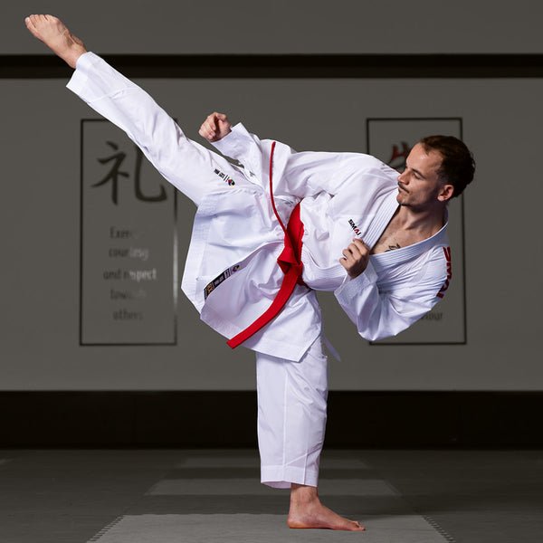 A martial artist executes a high side kick in a dojo, clad in the WKF Reversible Karate Uniform - 6oz Premium Kumite Gi - Inazuma RX by SMAI, complete with a red belt. The white gi contrasts strikingly against the backdrop of calligraphy-adorned walls.