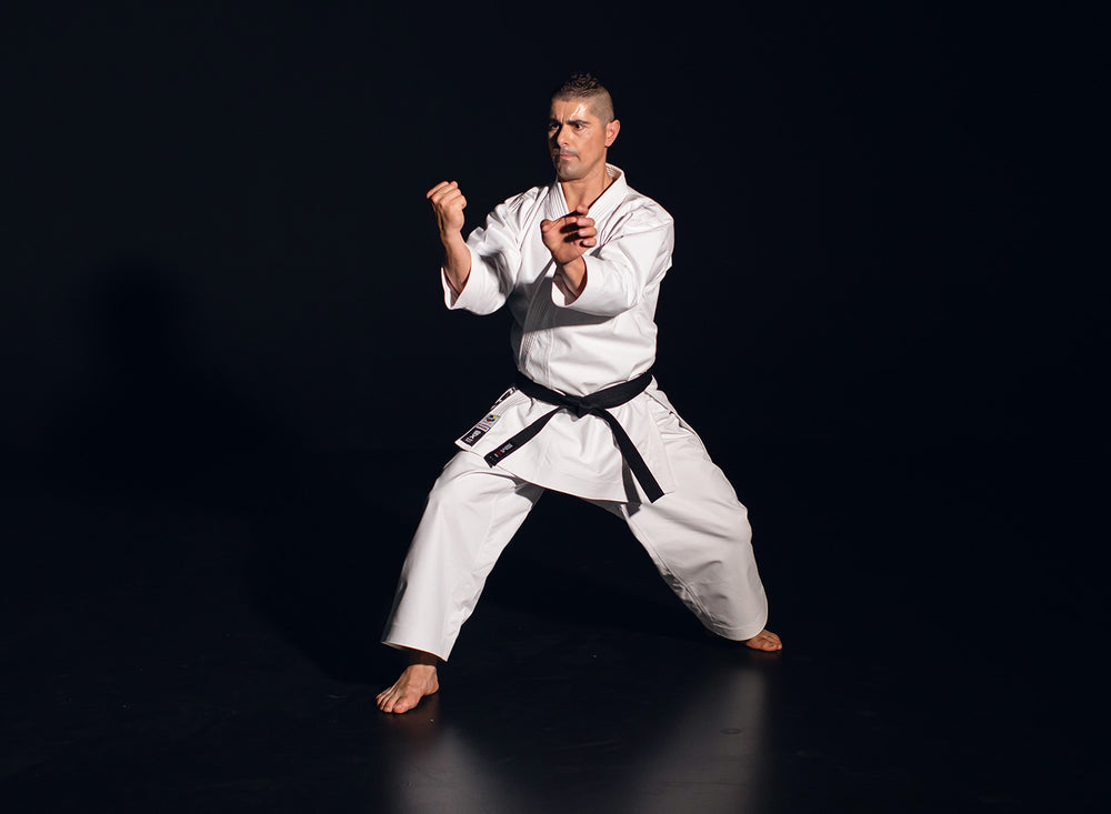 A man wearing a white karate gi and black belt stands barefoot in a martial arts pose against a dark background, with his fists raised and legs apart in a defensive stance.