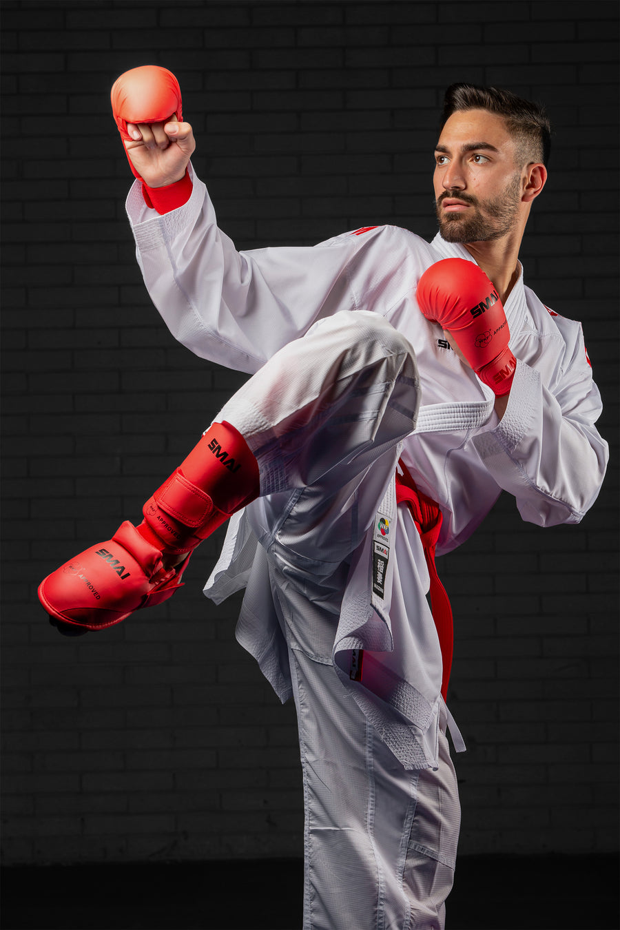 A male martial artist in a white uniform and red gloves performs a high kick, looking focused and poised against a dark brick background.