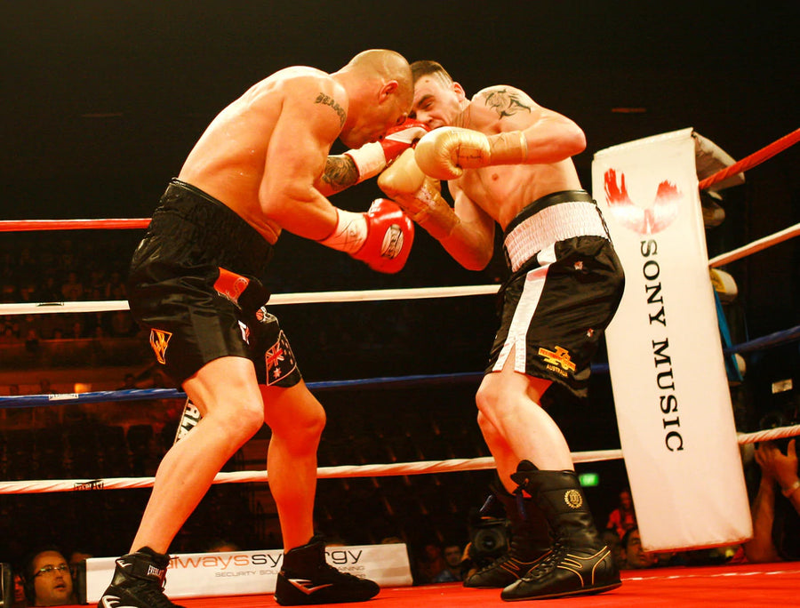 Two boxers in a ring, wearing shorts and gloves, engage in an intense punch exchange. The ring features branding such as Sony Music. The audience is visible in the background.