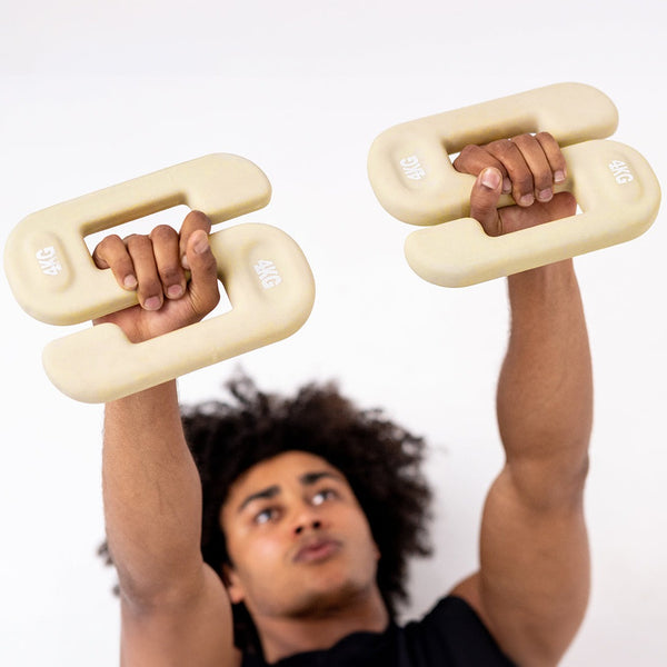 In the midst of a bustling commercial gym, an individual is performing free weight training with two SBELL Chalk 4 kg plates. These distinctively designed, beige weights feature built-in handles for a secure grip, highlighting an intense concentration on hand movement and precision as the weights are lifted.