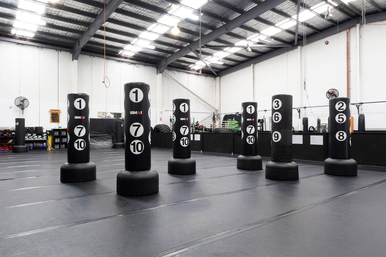 A spacious martial arts training gym with eight black standing punching bags, each labeled with numbers, arranged in two rows on a padded floor under a high ceiling with exposed beams.