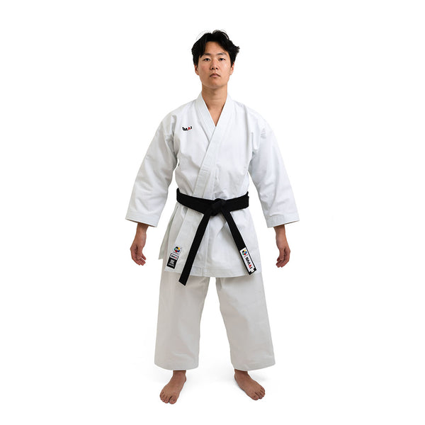 A martial artist in a white SMAI WKF Karate Uniform - Premium Kata Gi - Arashi stands barefoot on a clean backdrop, proudly displaying their black belt and patches on the left side and belt, embodying discipline and focus with a neutral expression.