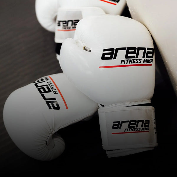 Close-up of white boxing gloves with "arena FITNESS MMA" written on them, lying on a dark surface next to a white wall.