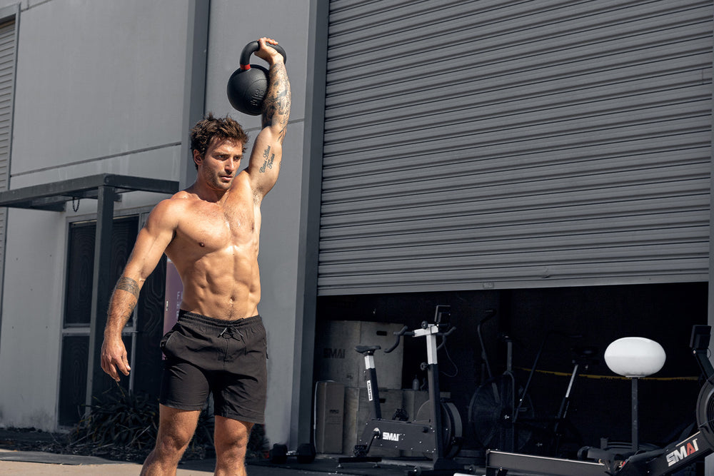 A shirtless man wearing black shorts lifts a kettlebell overhead with one arm outside an industrial building, showcasing his muscular physique under bright sunlight. Fitness equipment is visible in the background.