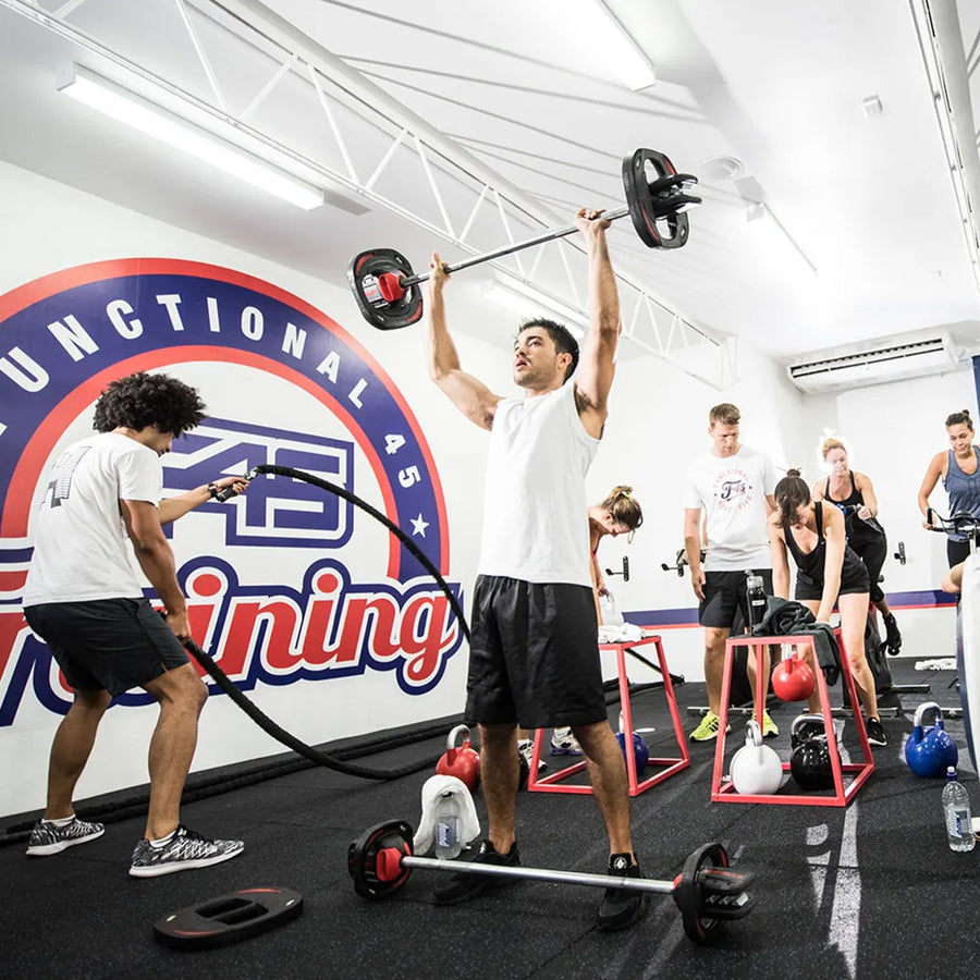 A group of people exercise in a gym, lifting weights and using equipment. One man lifts a barbell overhead in the foreground. The wall has a large "F45 Training" logo. Others work out in the background.