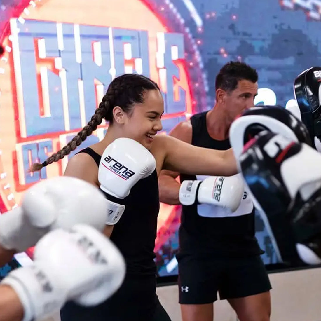 People are engaged in a boxing class, wearing gloves and focused on training. A woman with braided hair is smiling, and a man is visible in the background. A large, colorful sign is partially visible behind them.