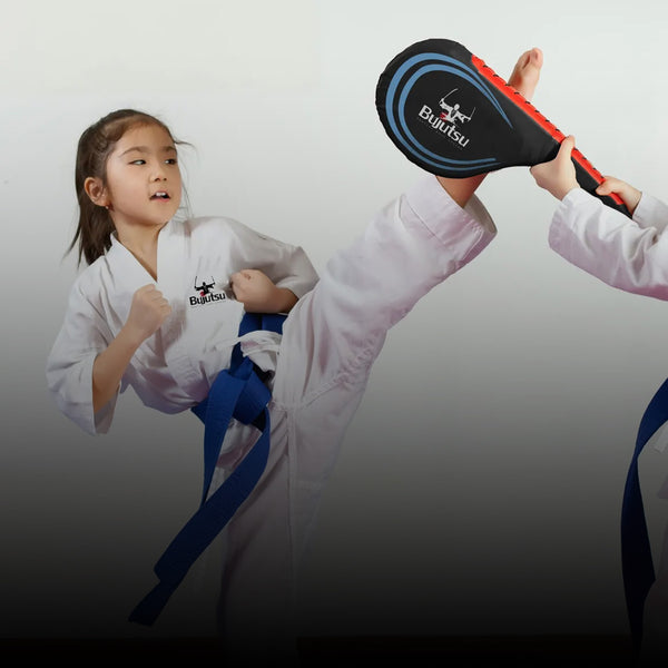 A young girl in a martial arts uniform executes a high kick towards a padded target held by another person. She wears a blue belt and appears focused on her practice.