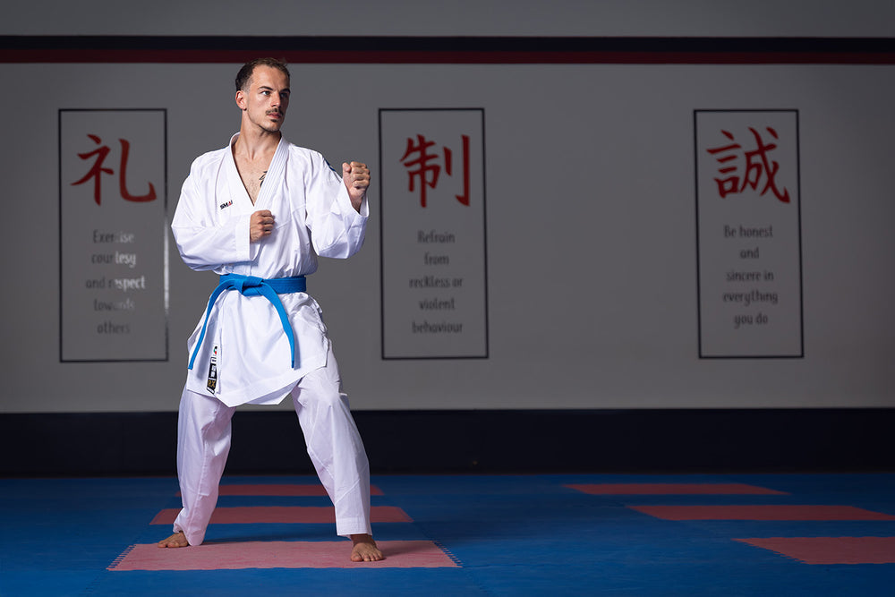 A man in a white karate uniform with a blue belt stands in a martial arts stance on a mat. Behind him, three wall posters display Japanese characters and positive motivational phrases.