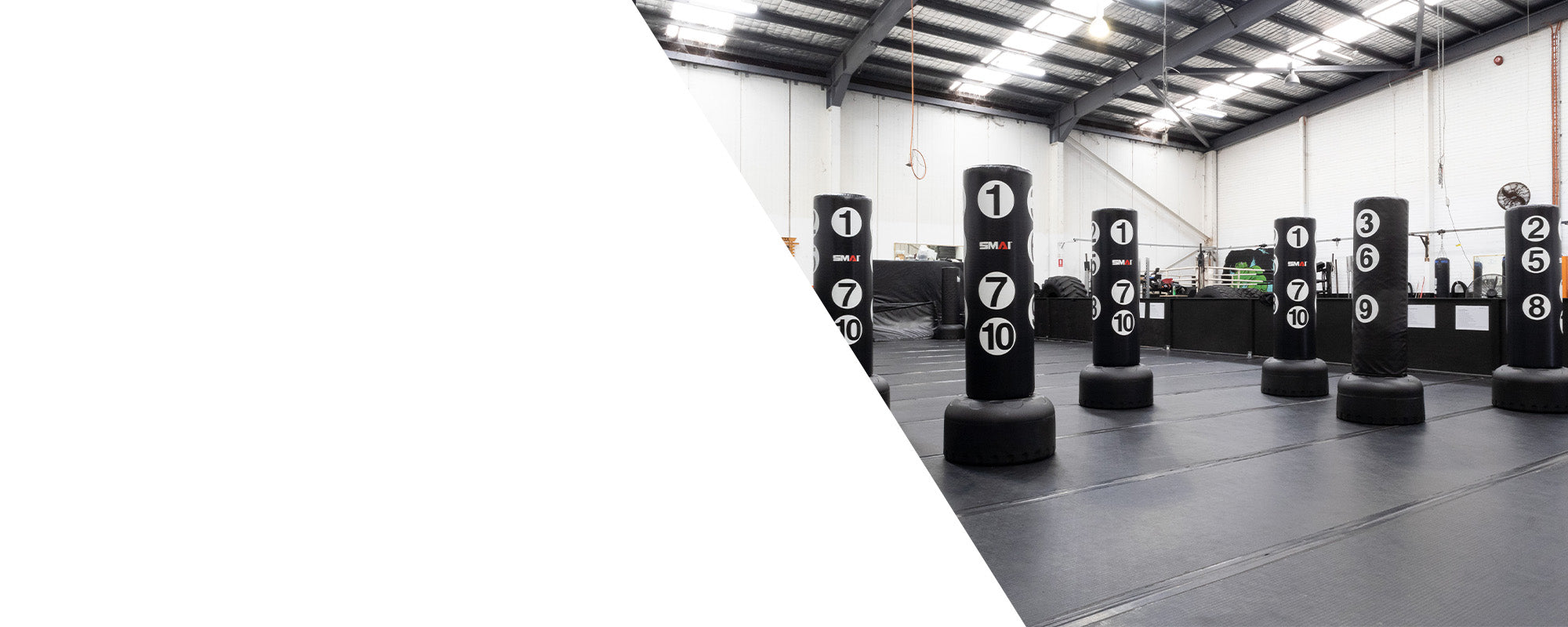 Several numbered punching bags arranged in rows inside a spacious, well-lit gymnasium with black flooring and a high ceiling; the left side of the image features a large white geometric shape.