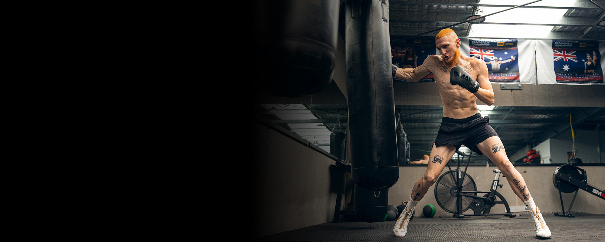A tattooed man with orange hair wearing boxing gloves, black shorts, and white shoes trains by punching a heavy bag in a gym with flags and exercise equipment in the background.