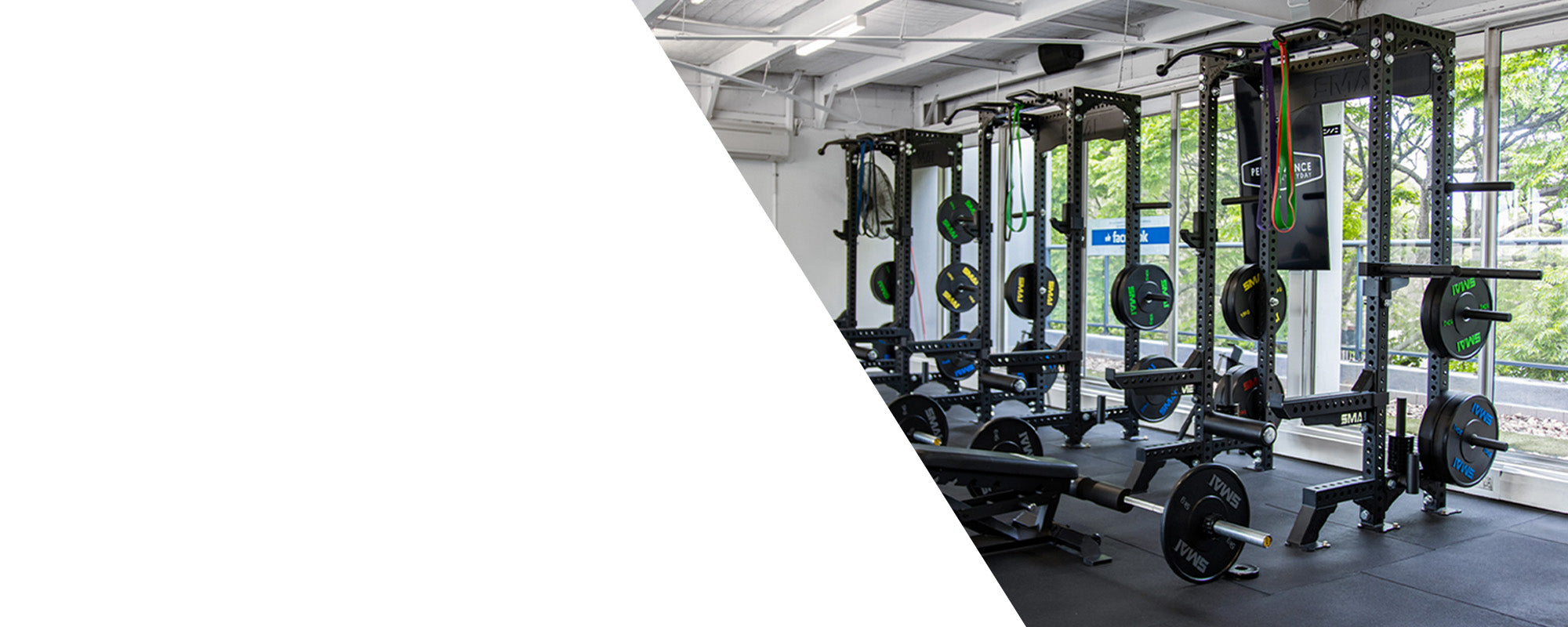 A modern gym with weightlifting racks, barbells, and benches next to large windows letting in natural light; the left side of the image is blank with a white background.