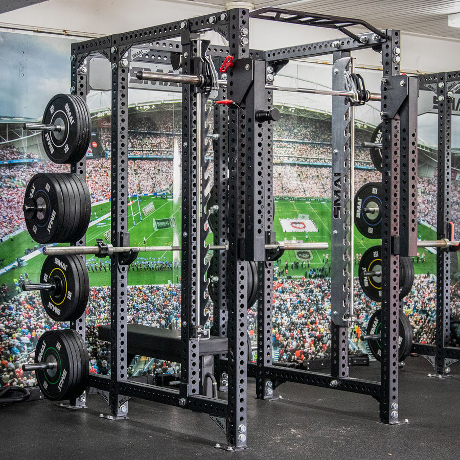 A heavy-duty squat rack loaded with weight plates stands in a gym. The background wall features a large mural of a crowded sports stadium and field.