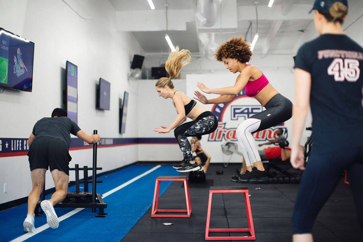 Four people are exercising in a gym. Two women are jumping over red hurdles, a man is pushing a weighted sled, and a trainer in black workout clothes watches. The gym has white walls and fitness equipment.
