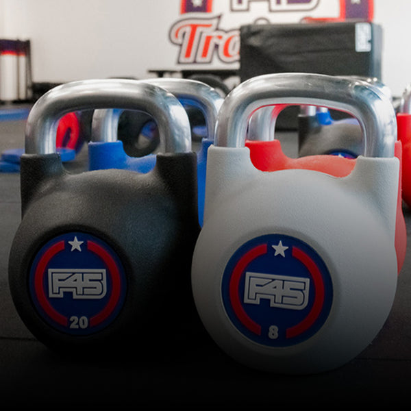 Two kettlebells with "F45" logos are placed on a gym floor. The black kettlebell is labeled "20" and the white one "8". Bright gym equipment and logos are visible in the background.