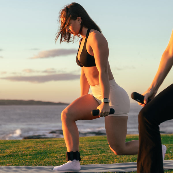 A woman in athletic wear lunges on a mat near the ocean at sunset, using SMAI Silicone Hand Weights - 1kg (Pair) with ergonomic grips; another person, partially visible, holds a dumbbell in the calm, scenic setting.