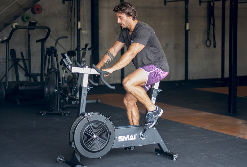 A man wearing a dark t-shirt and purple shorts rides an air bike in a gym with exercise equipment and rubber flooring visible in the background.