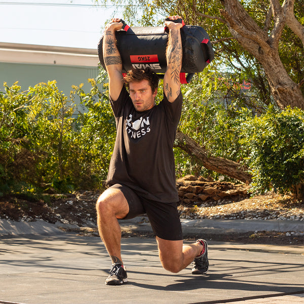 A man working out outdoors executes a dynamic lunge with a SMAI Core Bag lifted overhead. Clad entirely in black attire, he is set against a backdrop of trees and a building, symbolizing strength and balance.