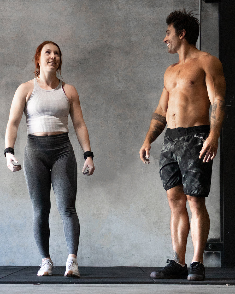 A woman in a white tank top and gray leggings stands next to a shirtless man in black shorts. They are both smiling, standing in front of a plain wall in a gym setting.