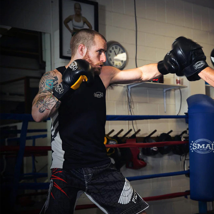 A man wearing boxing gloves and athletic gear throws a punch in a boxing gym, facing an opponent whose gloves are visible. Boxing equipment and a clock are in the background.