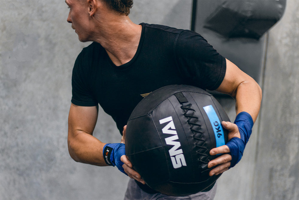 A person wearing a black shirt and blue wrist wraps holds a black 9kg medicine ball with both hands in a gym setting, standing near a concrete wall.