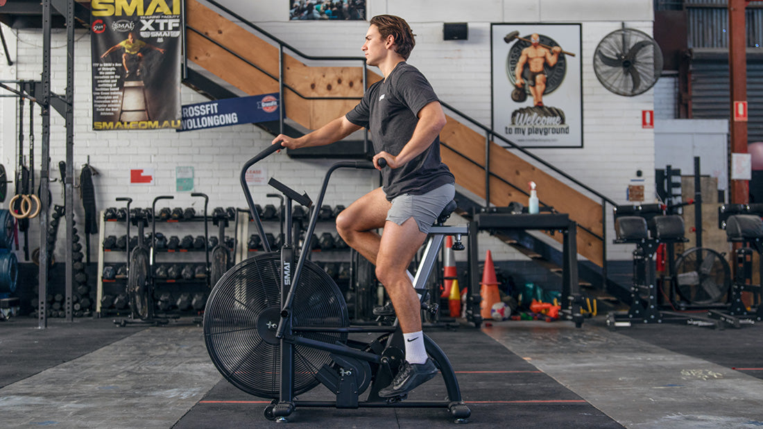 A man in athletic clothes rides an air bike in a gym with weights, equipment, and fitness posters visible in the background.