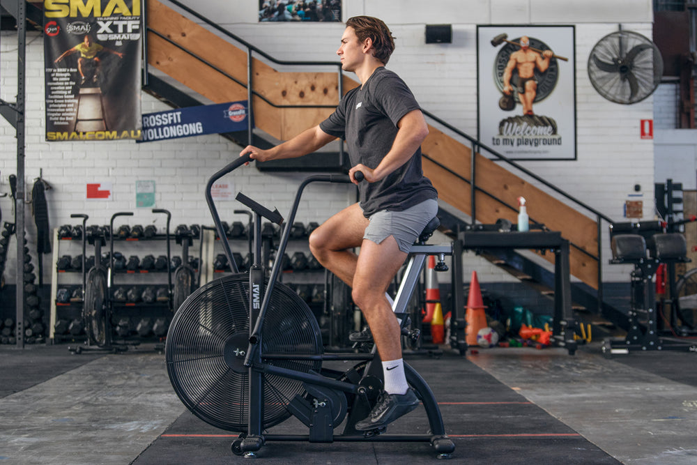 A person in athletic wear uses an air bike in a spacious gym with fitness equipment, posters, and wooden stairs in the background.