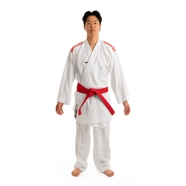 A person stands on a white background wearing the WKF Reversible Karate Uniform by SMAI, specifically the 6oz Premium Kumite Gi known as Inazuma RX, paired with a red belt. The gi features striking red logos on the shoulders that highlight focus and discipline. With their arms relaxed at their sides, they embody the true spirit of martial arts mastery.