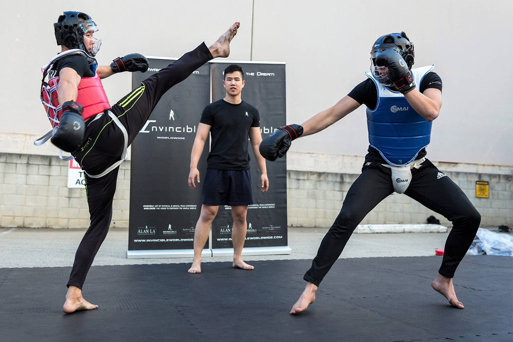 Two martial artists in protective gear spar on a mat, with one kicking high toward the other's head. A referee stands between them, watching the match closely. A banner and wall form the background.