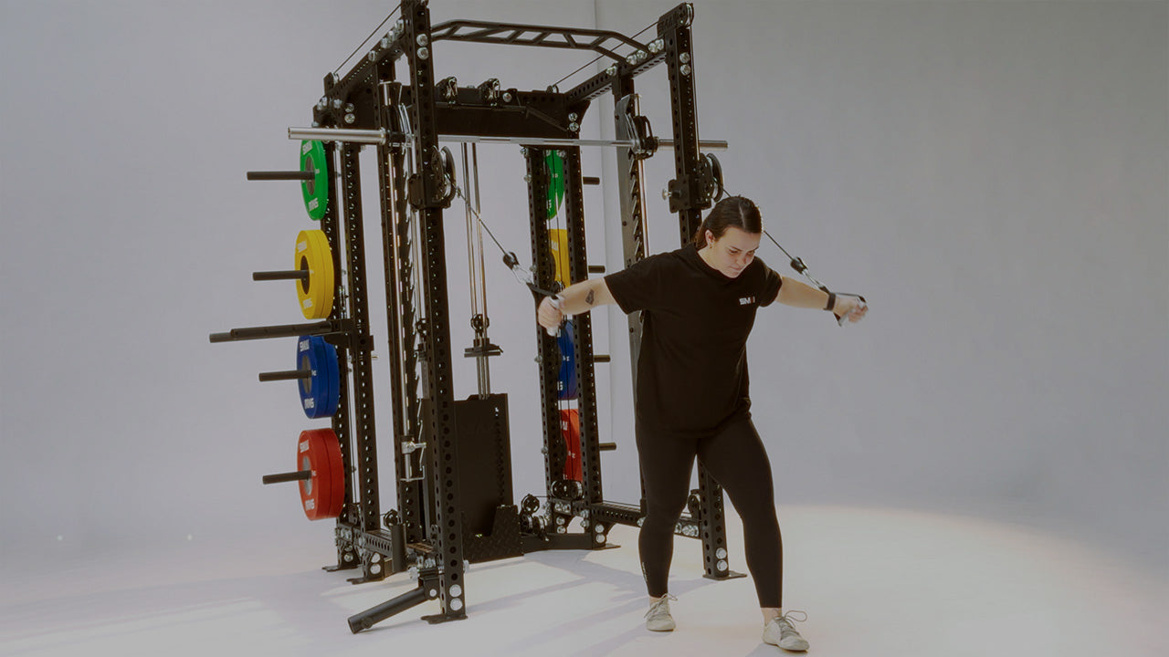 A person in black workout clothes uses a cable machine for exercise, pulling the handles forward with both hands; colored weight plates are visible on the machine.