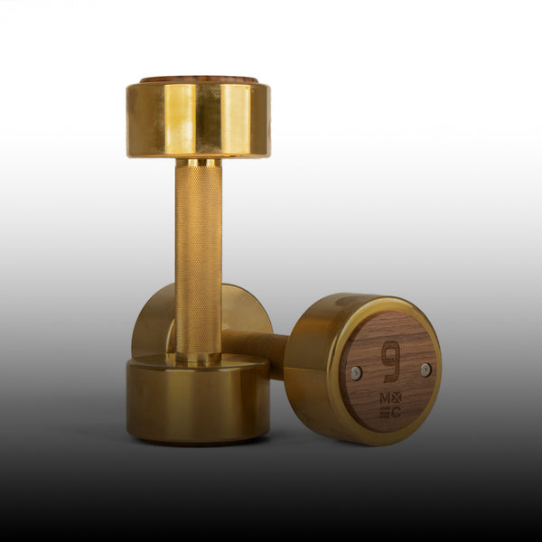 A pair of stylish, gold-colored dumbbells with a wooden accent on the ends. The number "9" and "MX EC" are engraved on the wooden part, suggesting the weight measurement. The background fades from white to black.