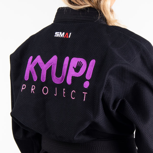 Person wearing a black martial arts gi with "KYUP! Project" embroidered in purple and pink on the back. The gi also includes an "SMAI" logo near the neck. Only the back and partial profile of the person are visible.
