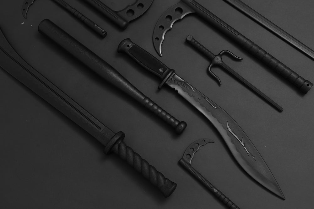 Several black bladed weapons and martial arts tools, including machetes, a large knife with a curved blade, a bat, and a sai, are arranged on a dark background.