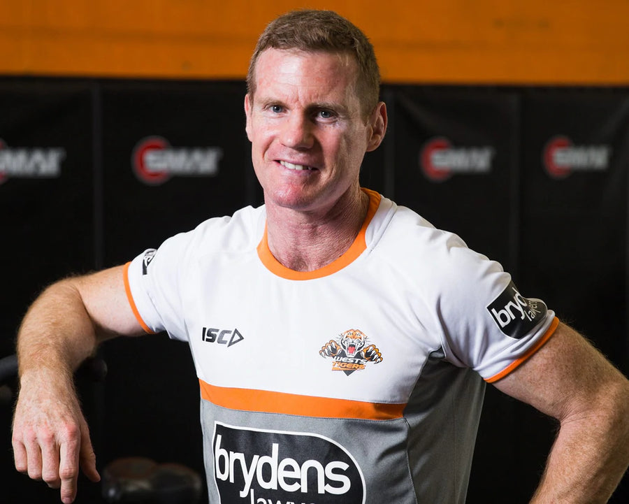 A man in a white and gray sports shirt with orange accents stands indoors. The shirt has logos and "Brydens" printed on it. He is smiling with his left hand resting on a fitness bike. The background is a gym setting.