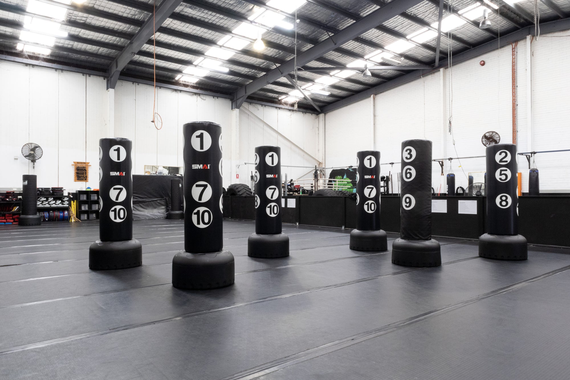 A gym with seven large standing punching bags, each marked with white numbers, arranged in two rows on black padded flooring under a high, open ceiling with exposed beams and lights.