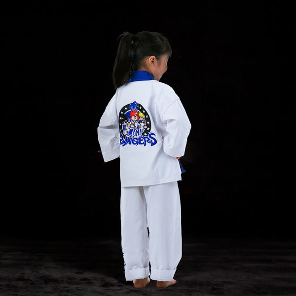 A young girl with a ponytail stands barefoot on a dark surface, wearing a white martial arts uniform with a blue collar. The back of her uniform features a colorful “Tiny Tigers” logo with a cartoon tiger.
