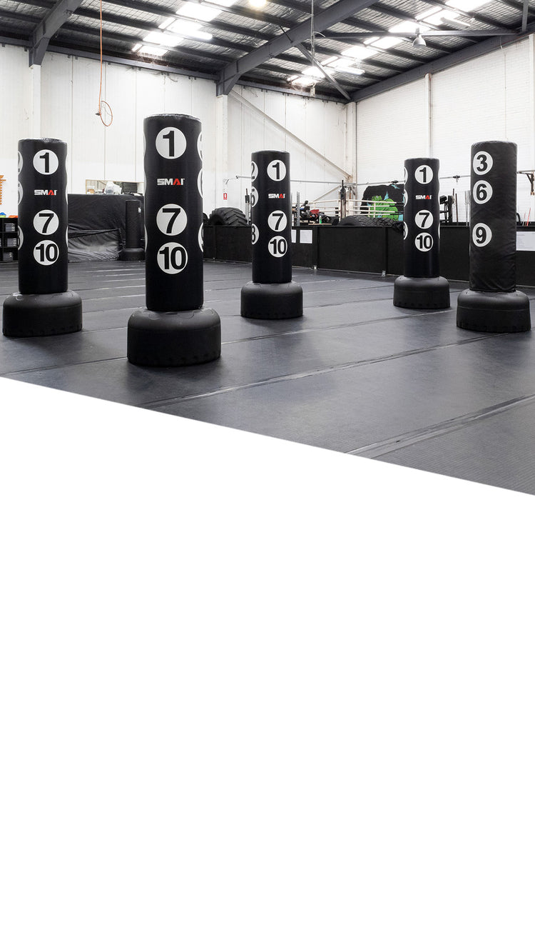 A spacious gym with several black standing punching bags, each labeled with white numbers 1, 7, and 10. The area has padded flooring and high ceilings with visible beams.