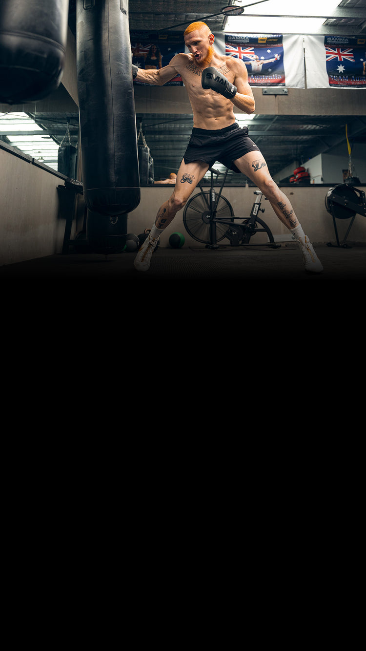 A shirtless man with tattoos and orange hair is kicking a heavy punching bag in a gym, wearing black shorts and white boxing shoes. Gym equipment and flags hang in the background.