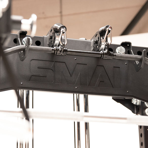 Close-up of the Cross Beam Logo Plate - Vanta Series fitness machine with "SMAI" featured on it. The image emphasizes its metal components and the logo plate, conveying a sense of durability and strength. The background is blurred to highlight the machine's robust structure.