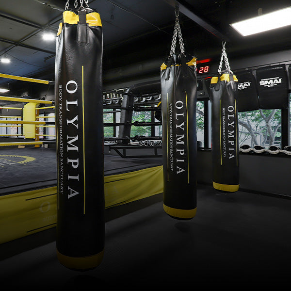 Three black and yellow punching bags labeled "Olympia" hang in a modern boxing gym, with a boxing ring and large windows in the background. The gym appears clean and well-lit, with various training equipment visible.