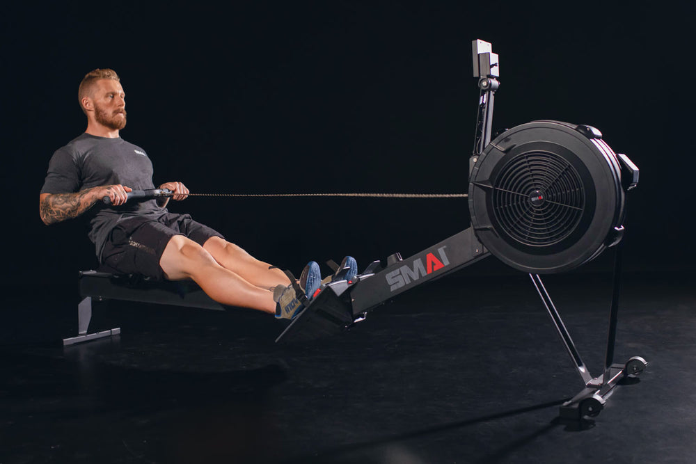 A man with tattoos is using a rowing machine in a dark room. He wears a grey t-shirt, black shorts, and blue shoes, focusing on his workout.