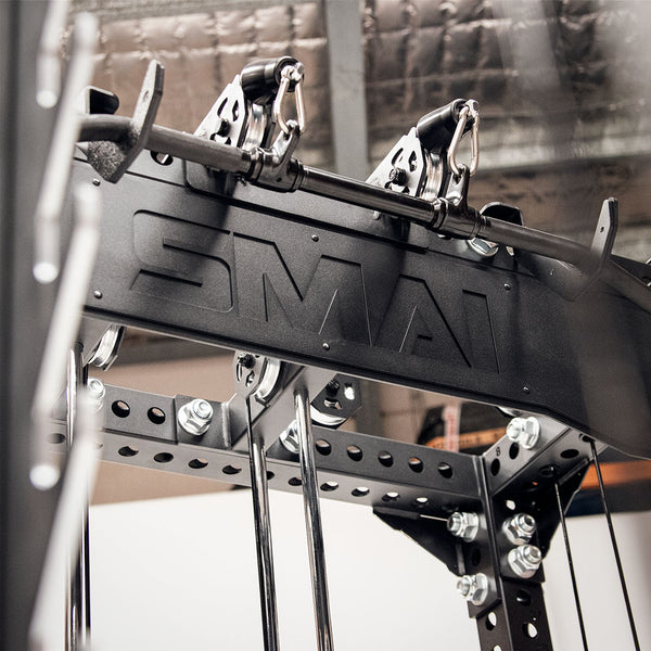 A close-up view of the Cross Beam Logo Plate from the Vanta Series showcases black gym pulleys with metal cables and handles. The structure is branded with "SMAI," set against an industrial-style ceiling background, emphasizing its robust design reminiscent of structural steel.