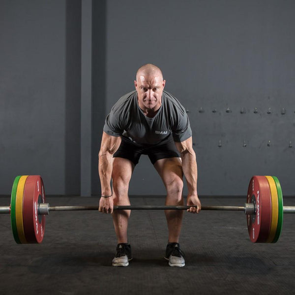 An individual dressed in athletic attire readies themselves to lift a barbell equipped with SMAI's Competition Bumper Plate 25kg (PAIR) - IWF Colour in a gym. The plain dark wall in the background amplifies their determined expression and stance, evoking the intense focus observed during weightlifting competitions.