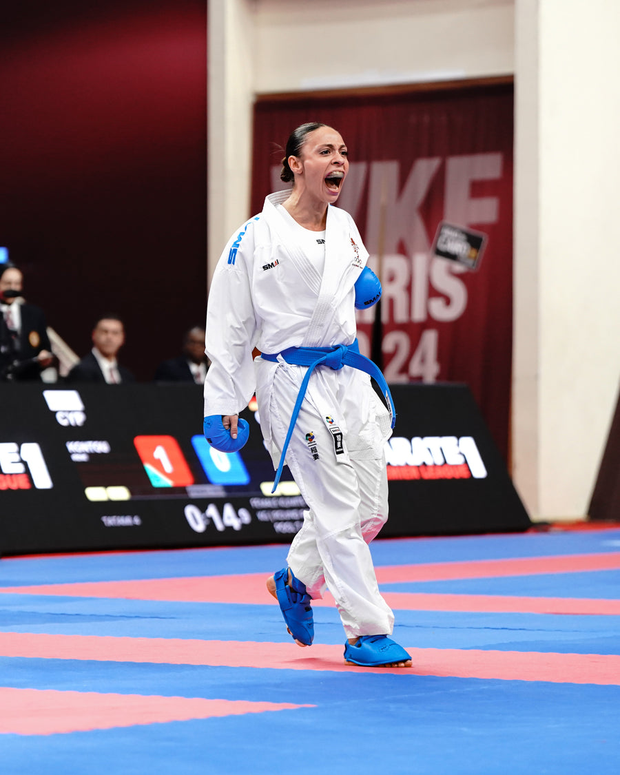 A female karate athlete in a white uniform and blue belt shouts passionately during a match on a blue and red mat, with referees and a digital scoreboard visible in the background.